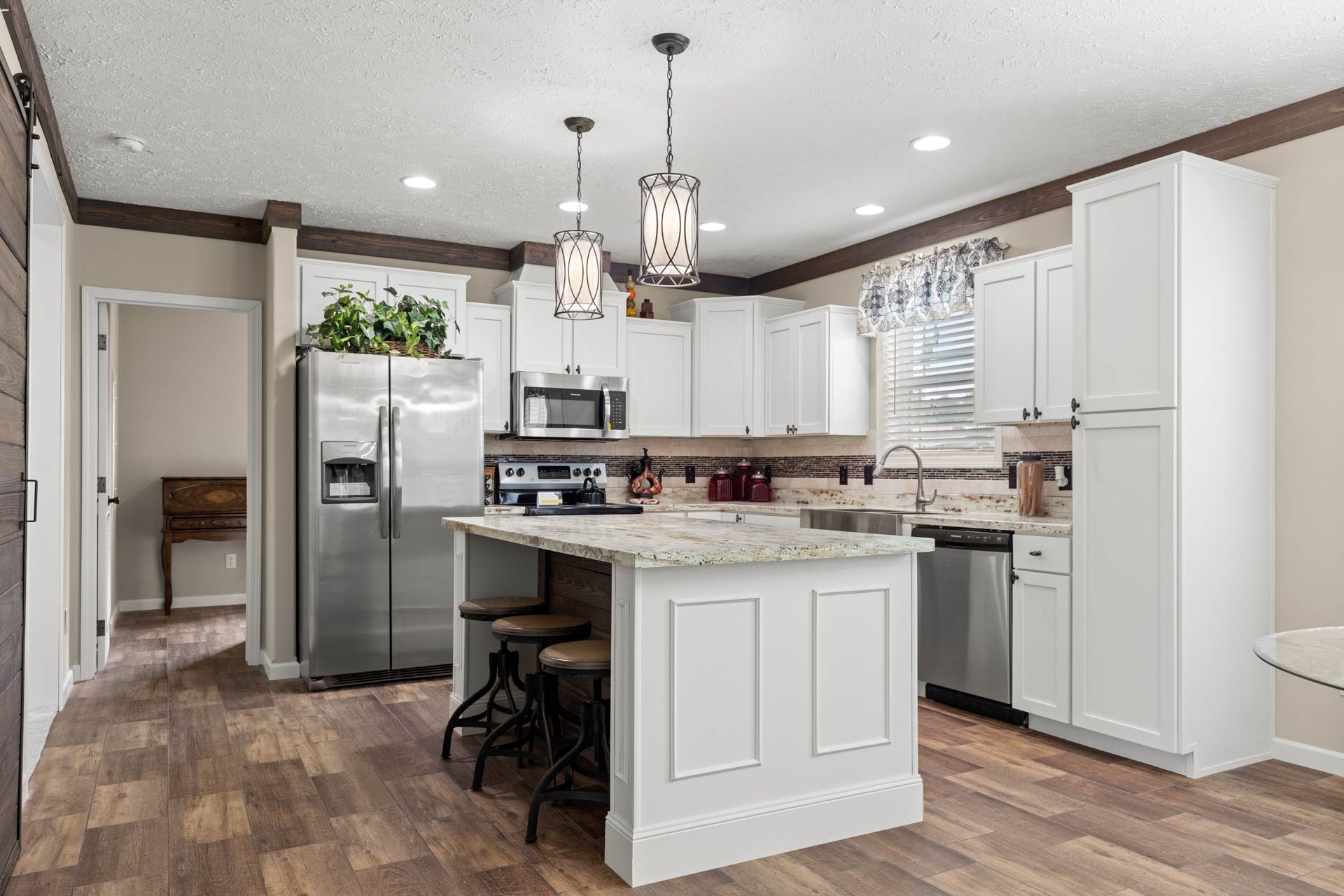 A modern kitchen featuring white cabinets, a stainless steel fridge, and appliances. An island with barstools sits under elegant pendant lights. Warm, inviting atmosphere.
