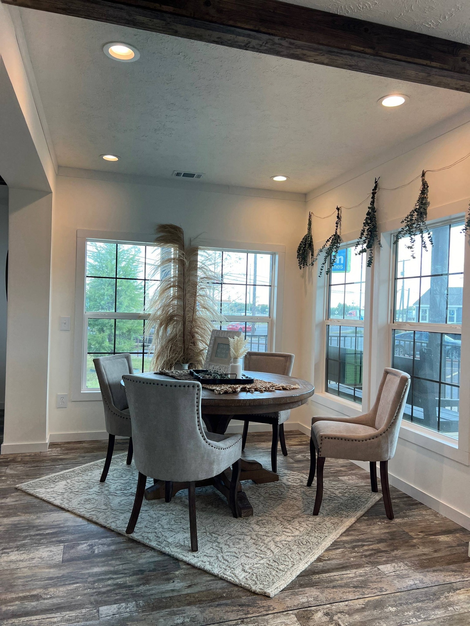 Cozy dining room with a round table, four beige chairs, dried pampas grass centerpiece, large windows, and hanging greenery, creating a serene ambiance.