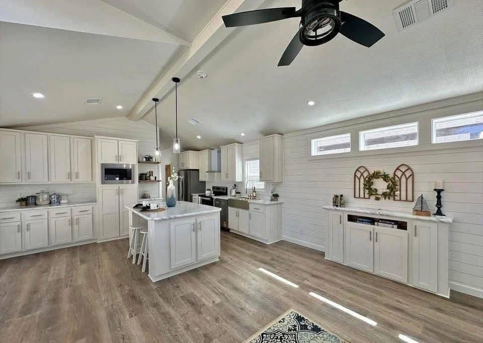 Spacious, modern kitchen with white cabinets, island, wood flooring, black ceiling fan, and decorative wreath. Bright, airy ambiance.