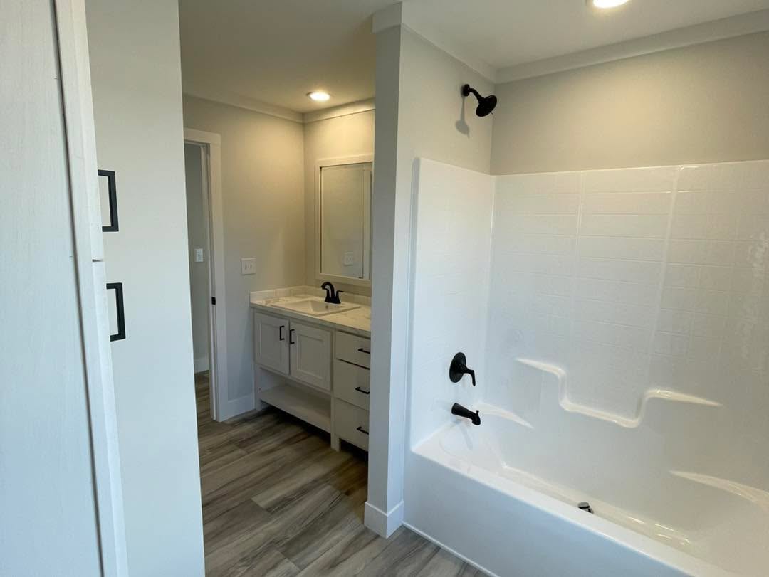 Modern bathroom with a large white bathtub, black faucet, and showerhead. Adjacent is a vanity with a mirror, white cabinets, and black handles.