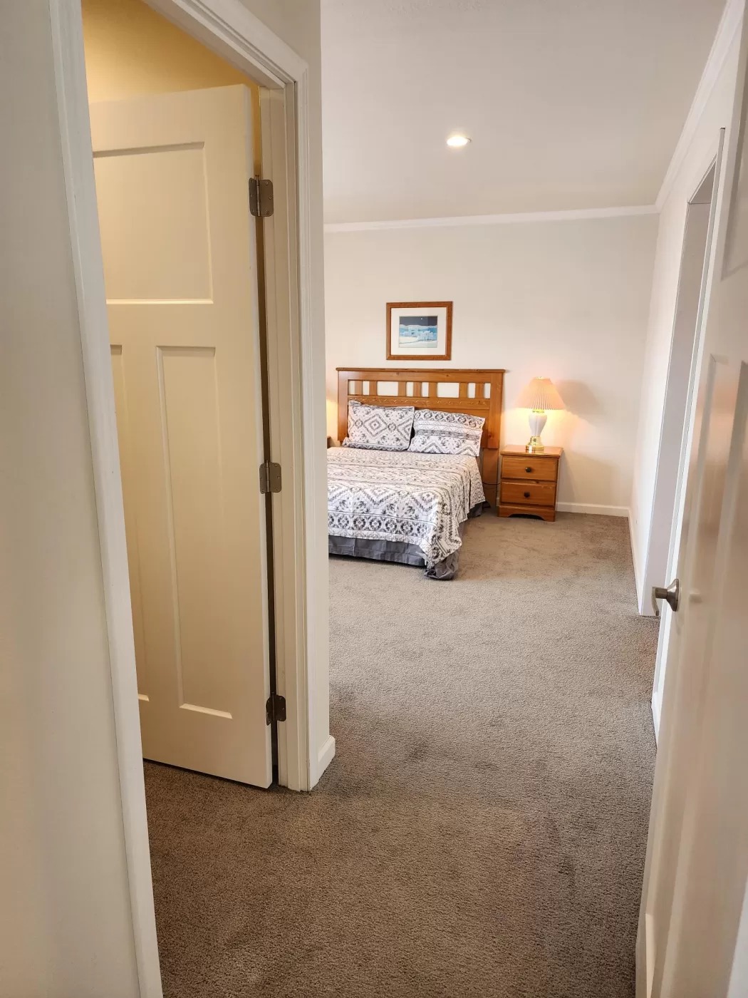 View into a cozy bedroom with a neatly made bed featuring patterned bedding, wooden headboard, and side table with a lamp. Warm, inviting atmosphere.