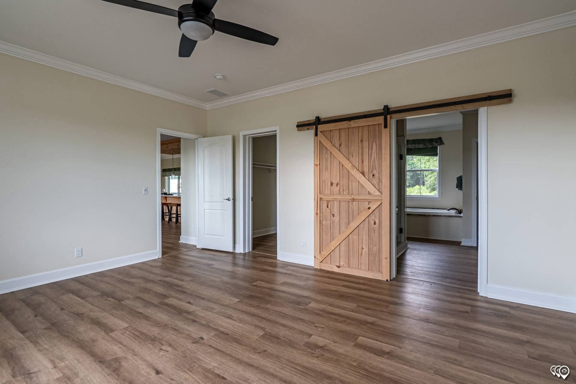 Spacious room with wood floor, ceiling fan, and cream walls. Features a sliding barn door revealing a bright room and white closet door. Calm tone.