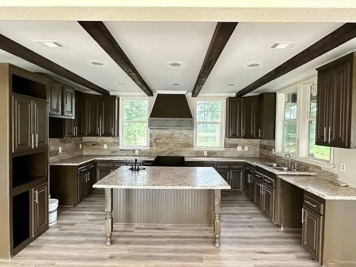 Spacious kitchen with brown cabinets and a large marble island. Dark wood beams contrast the light ceiling. Bright windows offer a view of greenery.