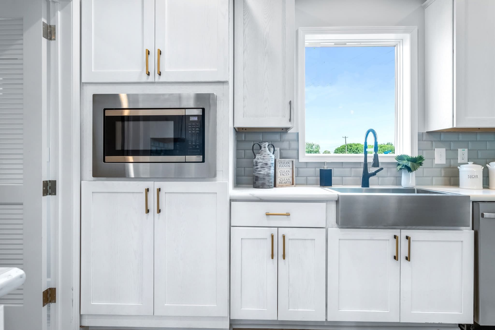 Bright kitchen with white cabinets and gold handles, featuring a built-in microwave and large stainless steel sink beneath a window with a blue sky view.