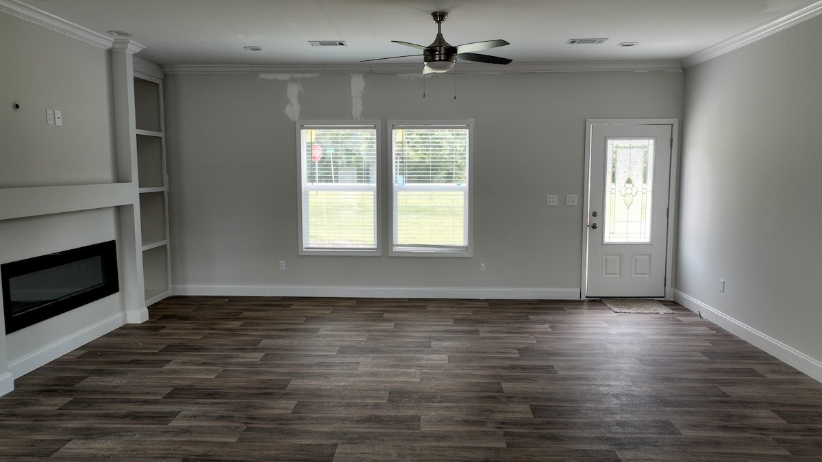 A spacious, empty living room with wood flooring, light gray walls, and a ceiling fan. Features include a modern fireplace, built-in shelves, and two large windows beside a white door, creating a bright and open atmosphere.