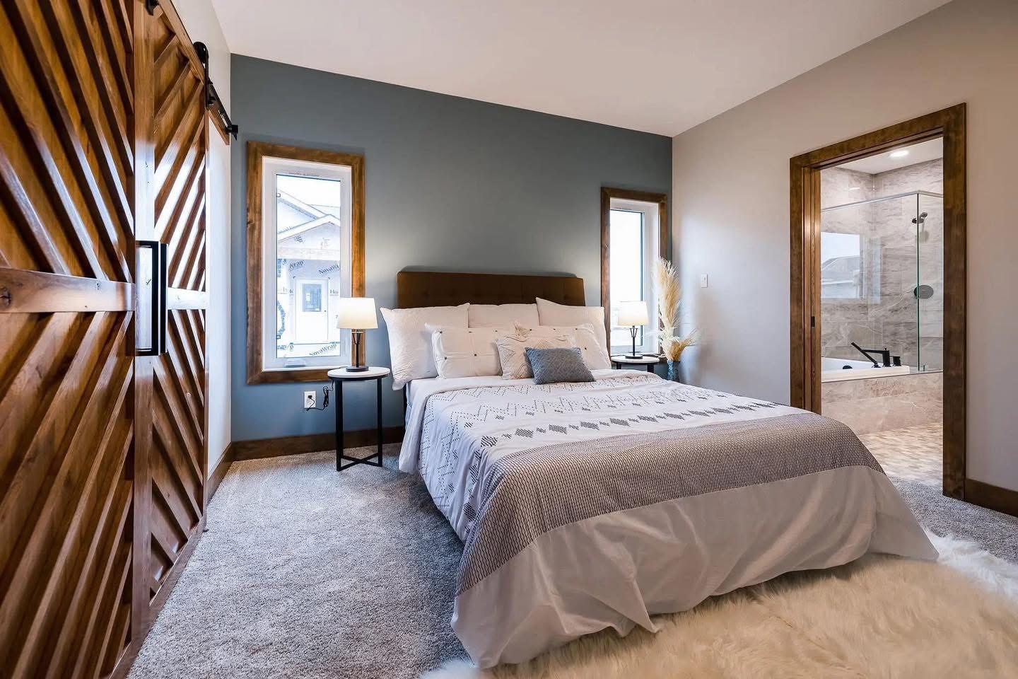 A cozy bedroom featuring a neatly made bed with white and gray bedding, a wooden sliding barn door, and adjacent en-suite bathroom with glass shower.