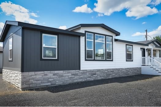 A modern prefabricated home with white and dark gray siding, large windows, and stone foundation. Bright sky with fluffy clouds overhead conveys a serene tone.