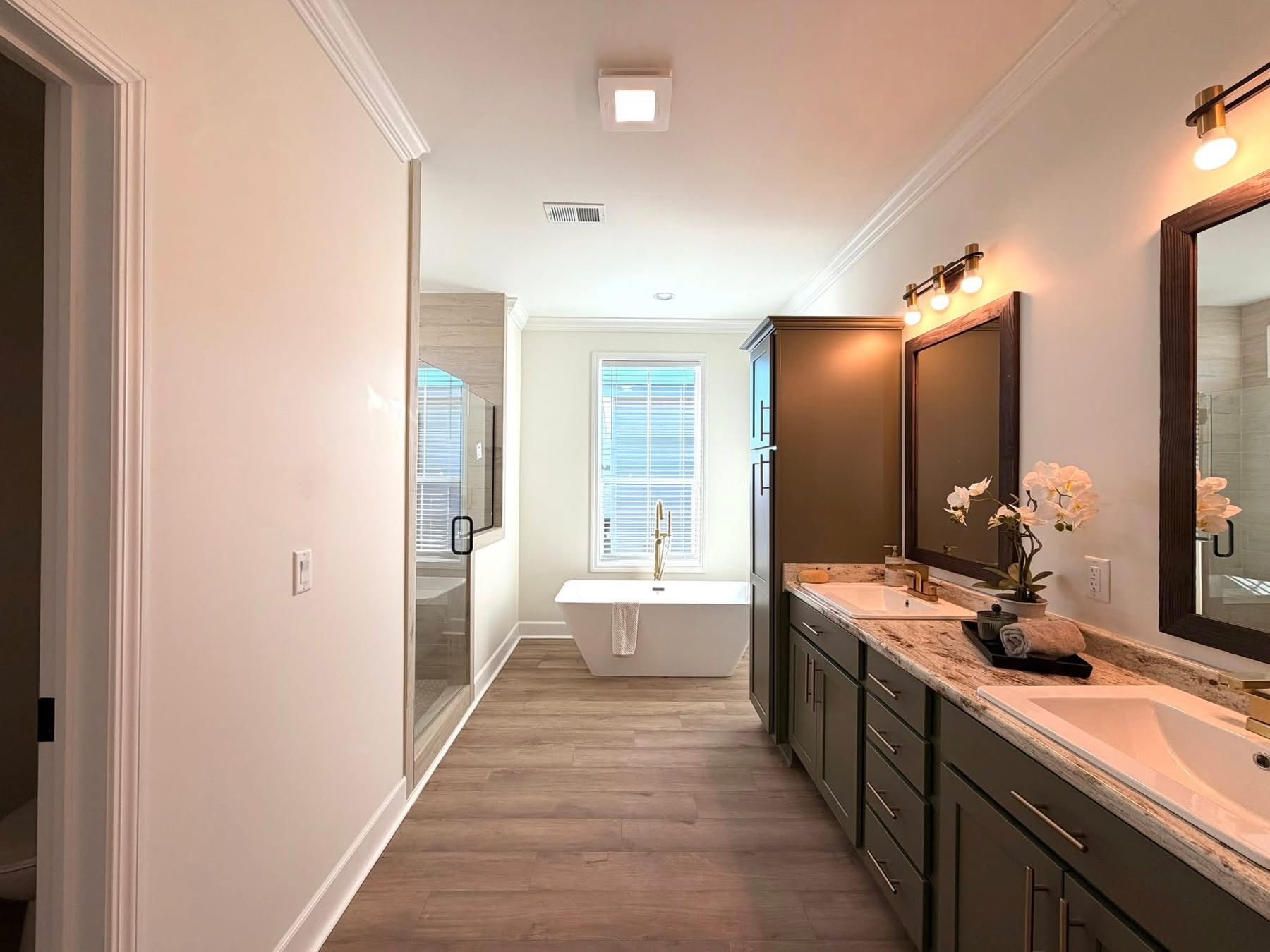 Bright bathroom with wood floors, freestanding tub beneath a window, and double vanity with green cabinets and mirrors. Modern, serene atmosphere.