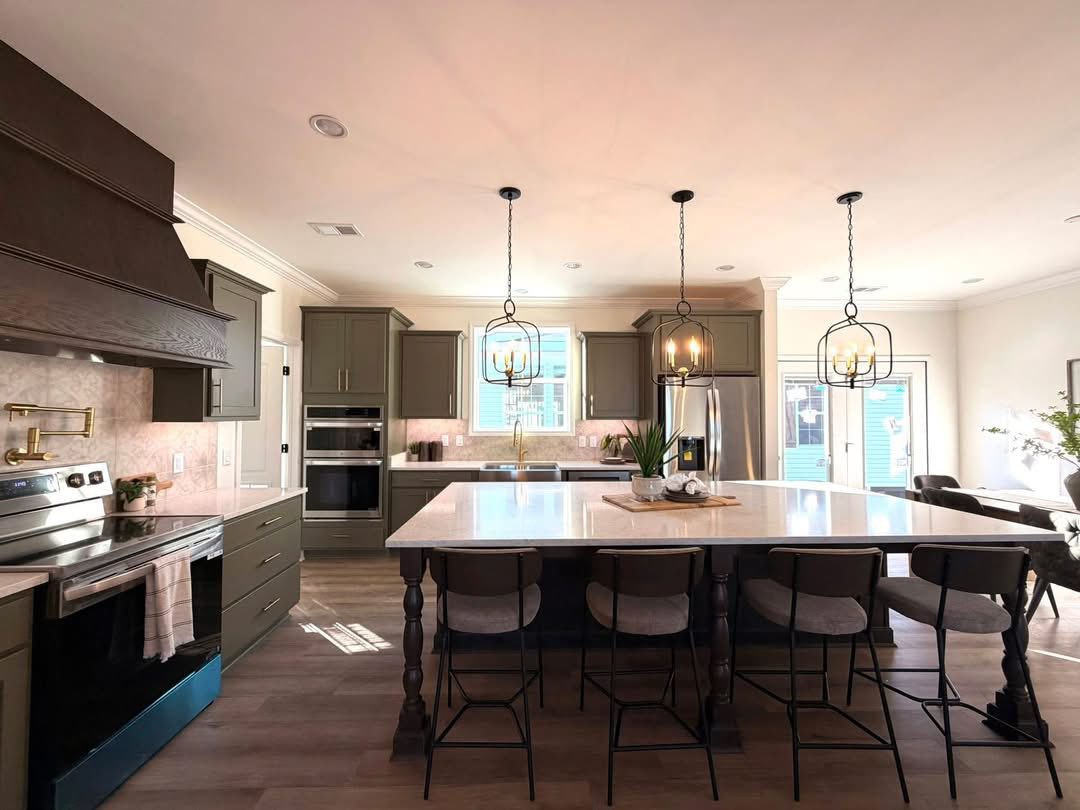 Stylish kitchen with a large island, five bar stools, pendant lights, and dark cabinets. Stainless steel appliances and sunlit wood floor convey warmth.