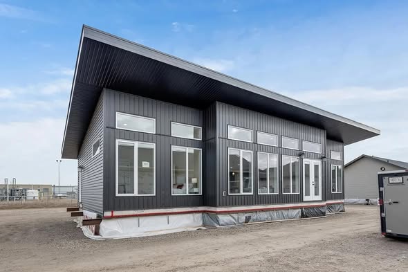 Modern single-story building with dark gray siding, large windows, and a slanted roof. Set on a construction site under a clear blue sky.