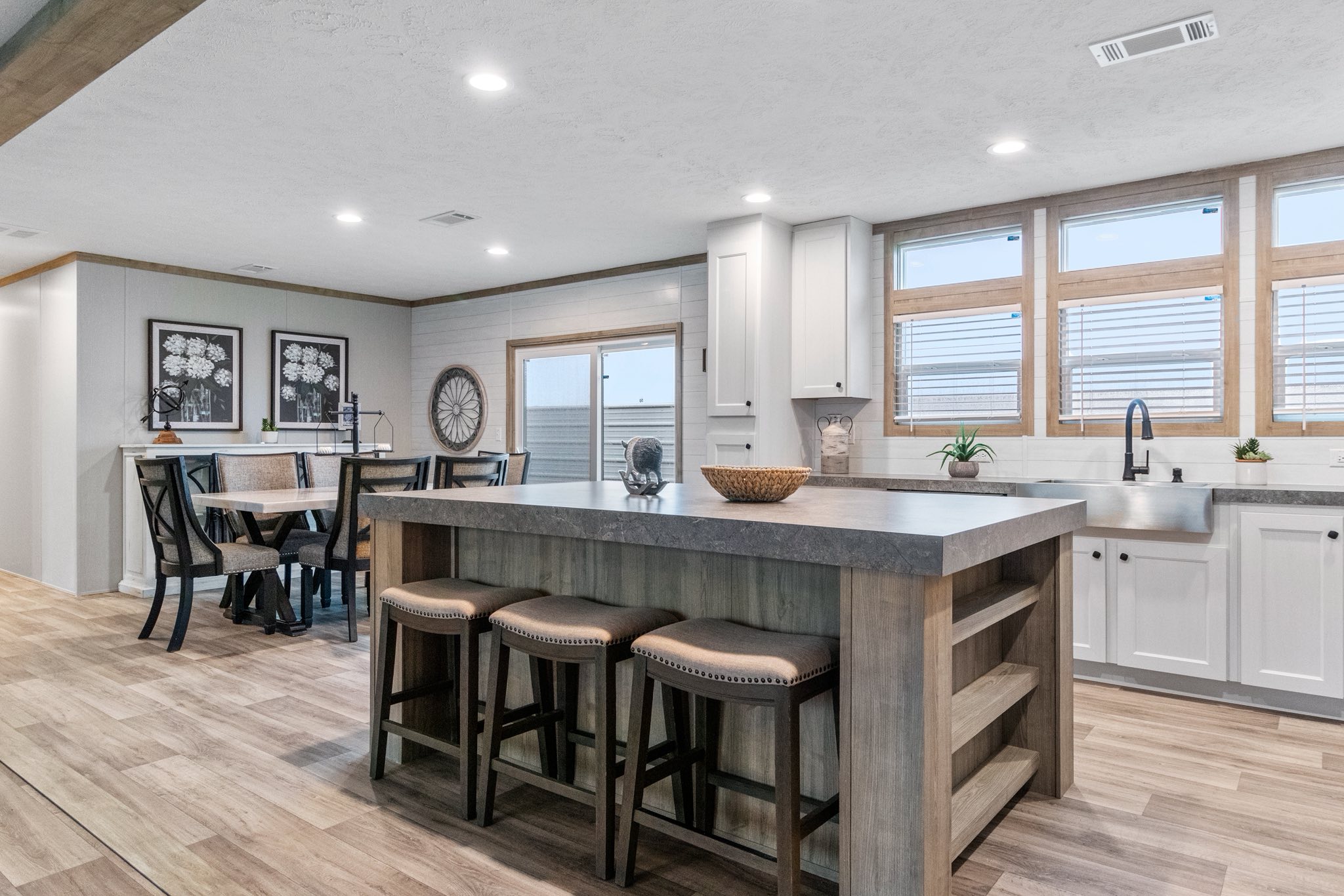 Modern kitchen and dining area with wooden floors. Features an island with stools, white cabinets, large windows, and a dining table. Bright and inviting.