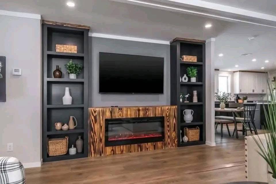 Contemporary living room with gray walls, a central mounted TV, sleek black shelves, decorative vases, plants, and a wood-accented electric fireplace.