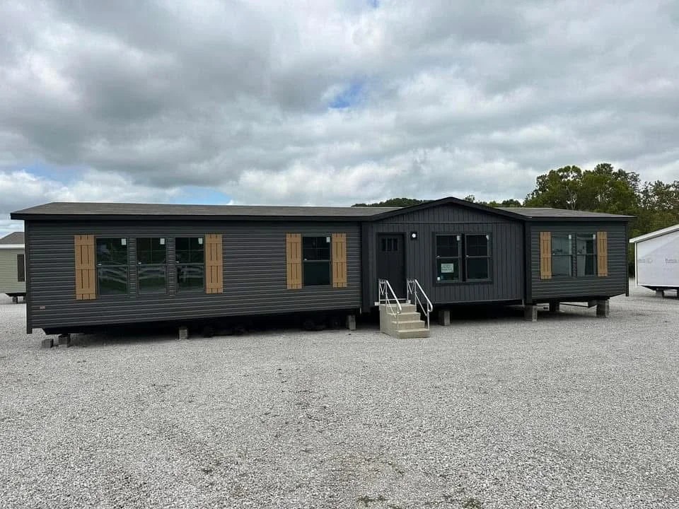A modern, dark gray manufactured home with wooden shutters and large windows sits on a gravel lot under a cloudy sky. Steps lead to the entrance.