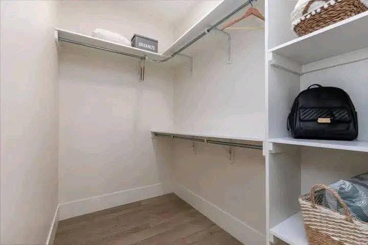 Minimalist walk-in closet with beige walls, wood floor, and metal rods. Shelves hold a black quilted bag, a basket, and an "organize" box, evoking a tidy, calm ambiance.