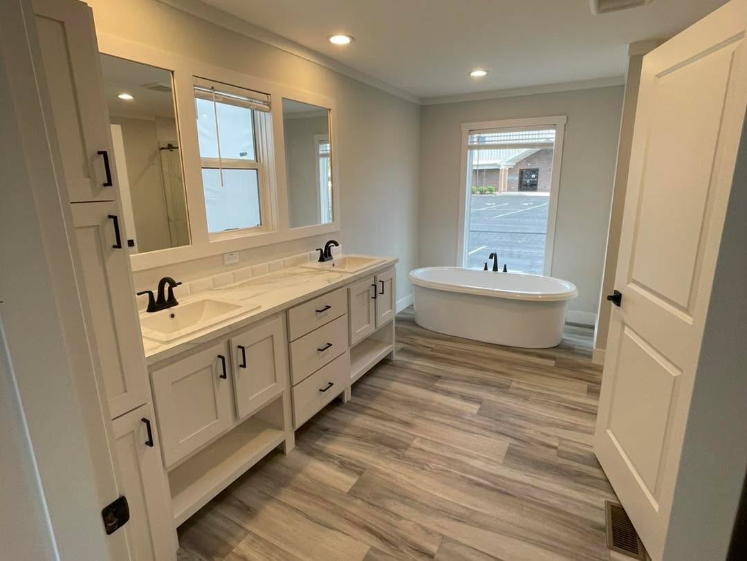 Spacious bathroom with a double-sink vanity and black fixtures on the left, a freestanding tub near a large window on the right, and wood-patterned flooring.