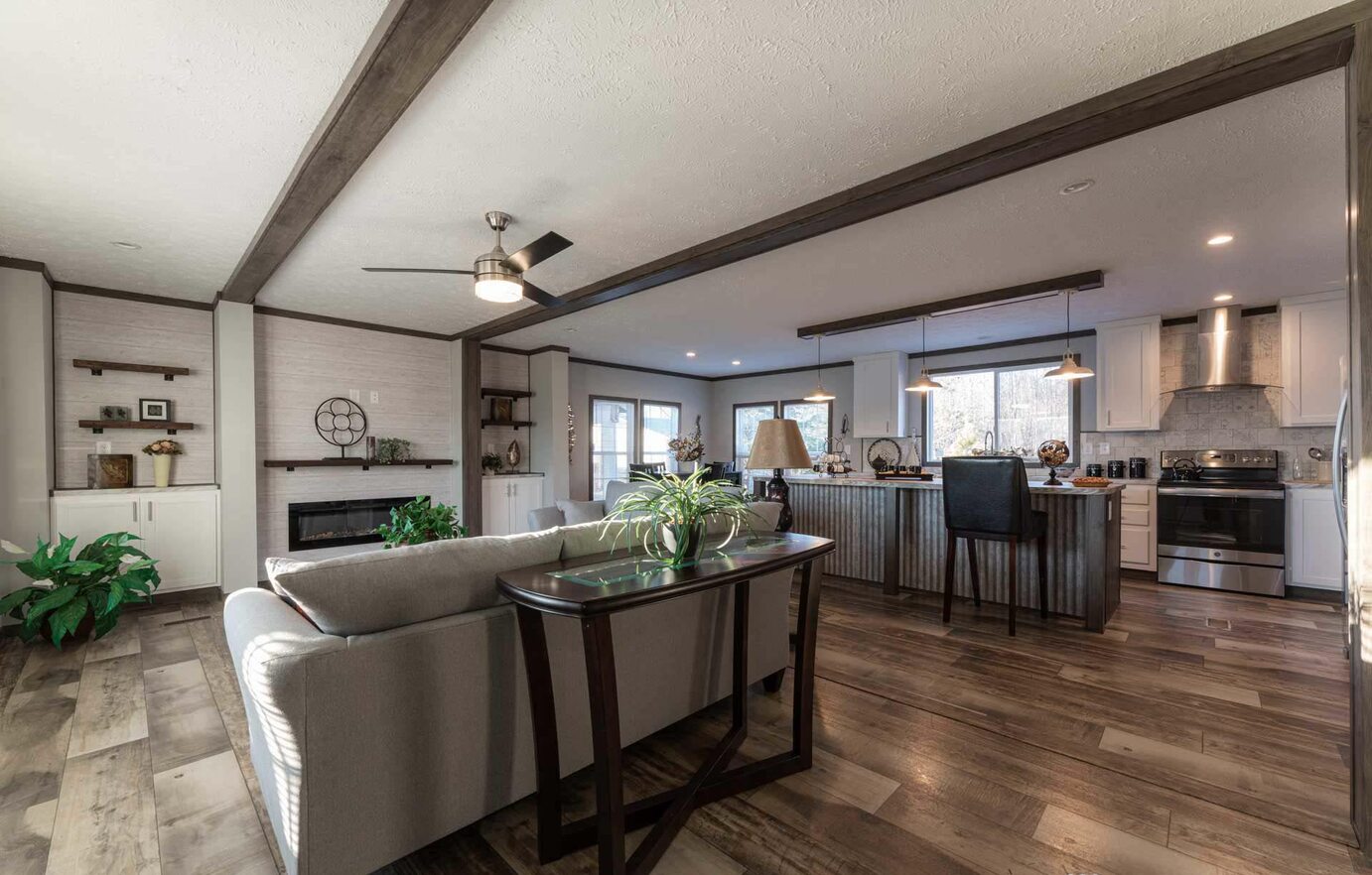 Modern open-concept living area with a cozy gray sofa, wooden flooring, ceiling fan, and kitchen island. Warm ambiance, neutral tones, and natural light.