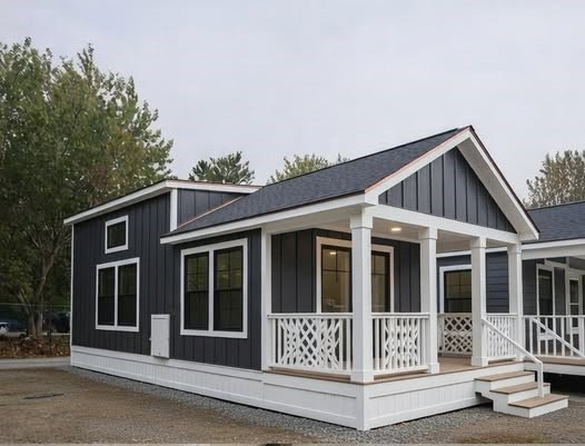 A small, modern gray house with white trim and a charming front porch is surrounded by trees. It conveys a cozy and welcoming atmosphere.