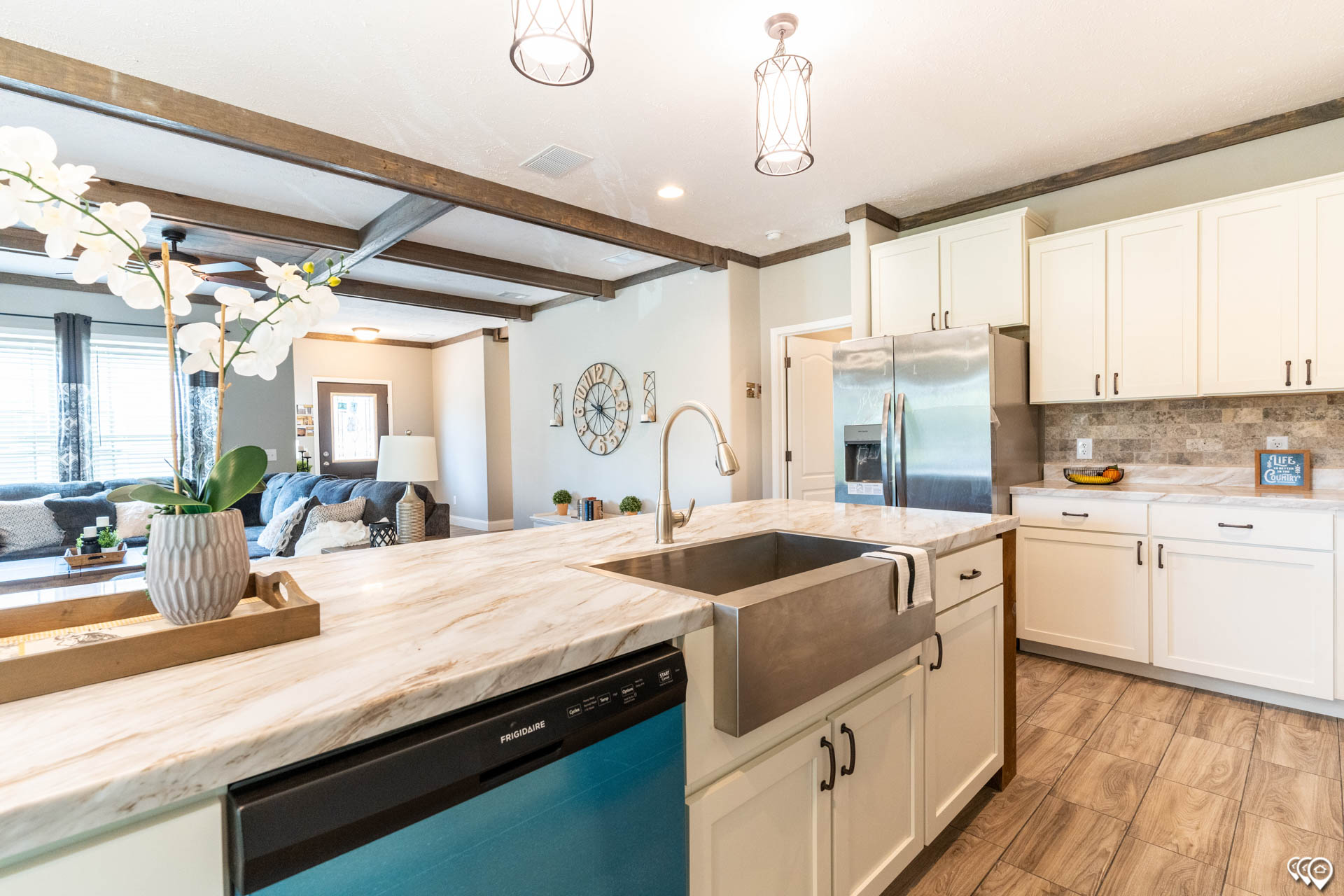 Spacious kitchen with white cabinets, marble countertops, and a farmhouse sink. Open layout with wooden beams, bright lighting, and cozy living area.