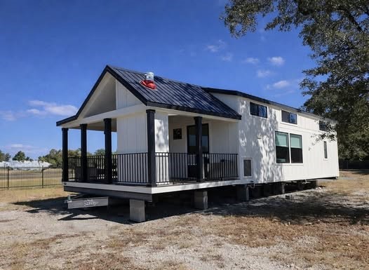 A modern tiny house with a pitched roof and white siding sits elevated on blocks. It features a small porch with black railings. The scene is sunny.