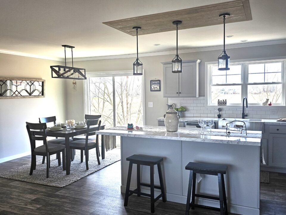 Modern kitchen with a marble island, gray cabinets, and pendant lights. Adjacent dining area has a dark wooden table and chairs, large window view.