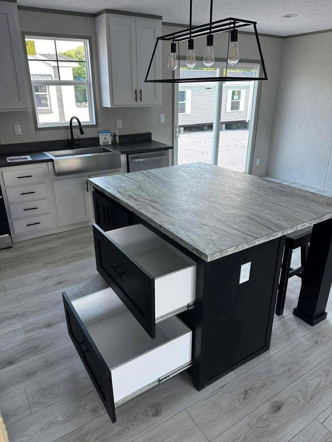 Modern kitchen features a central island with open drawers, light wood flooring, pendant lights, and sleek white cabinets near a large window.