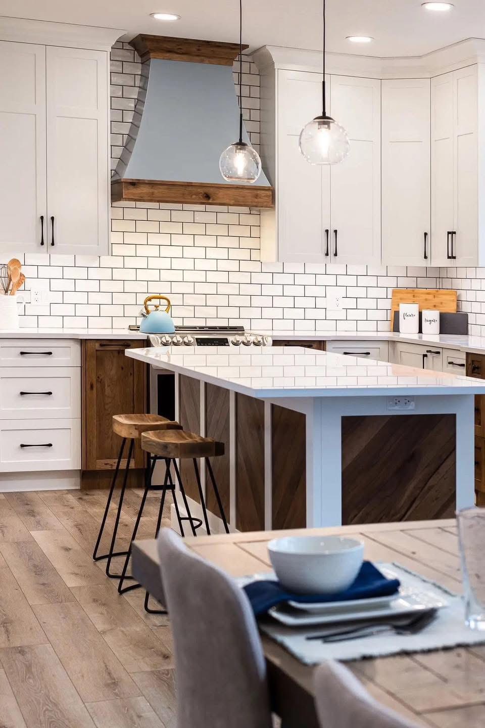 Modern kitchen with white cabinets, wooden accents, and a subway tile backsplash. Features a central island with wooden stools and pendant lights.