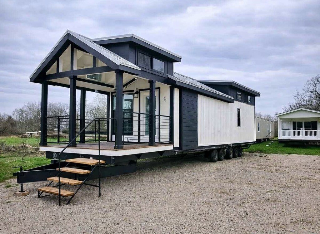 Modern tiny house on wheels with a dark and white exterior, an open porch, glass doors, and a gabled roof, set on a gravel space under cloudy skies.