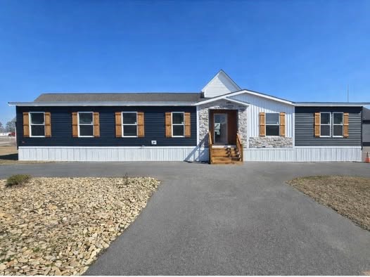 A modern manufactured home with blue siding, wooden shutters, and a small stone facade. It features a central gabled entrance and a paved driveway.