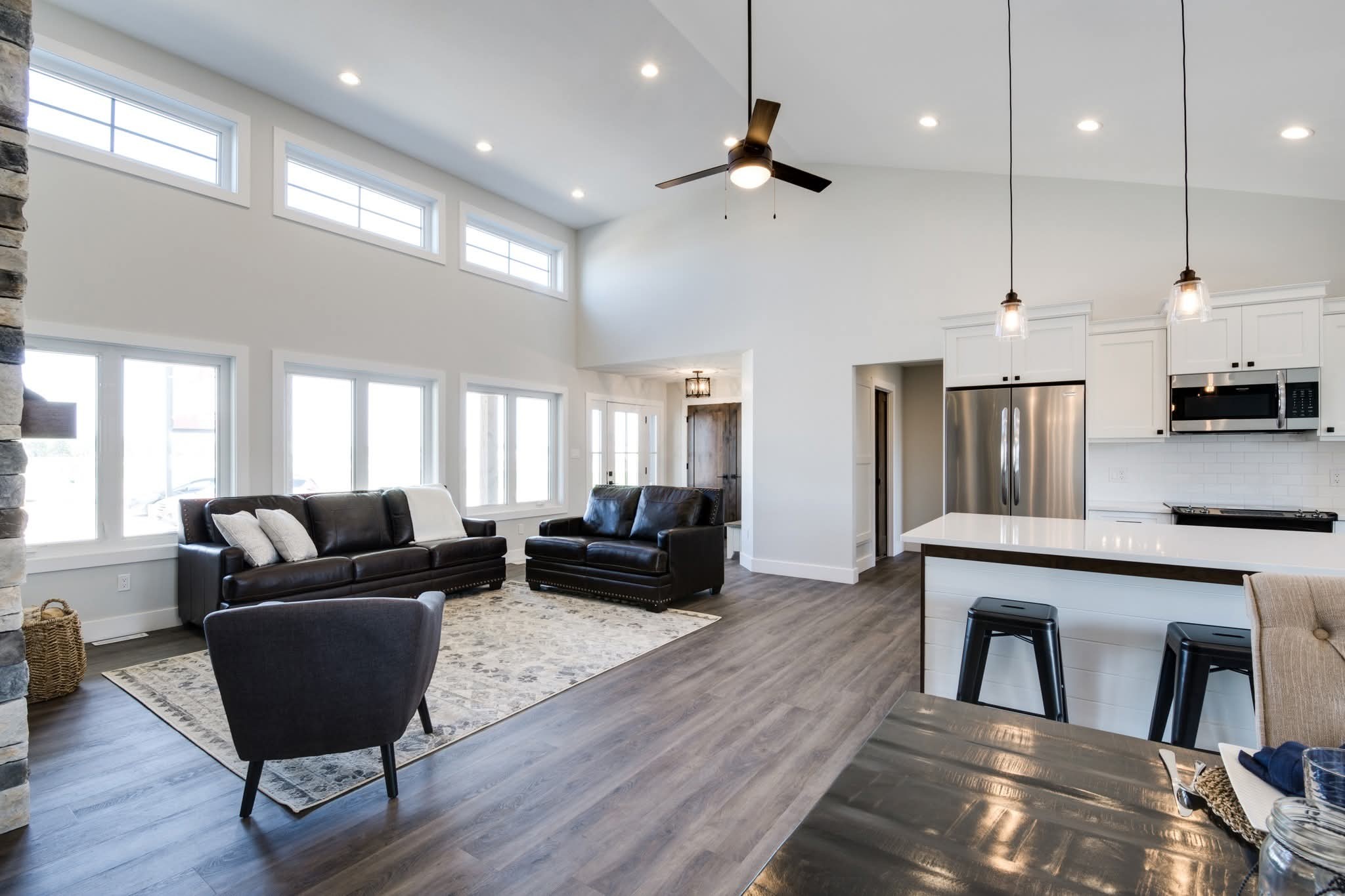 Spacious modern living room and kitchen with high ceilings. Features dark leather couches, a large rug, white cabinetry, and stainless steel appliances, creating an airy, elegant atmosphere.