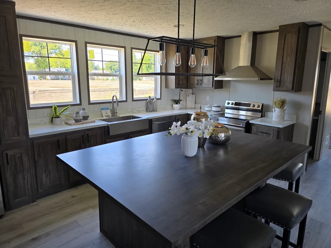 Modern kitchen with dark wood cabinets, large island with flowers, and industrial pendant lights. Sunlit windows overlook a grassy field. Cozy ambiance.