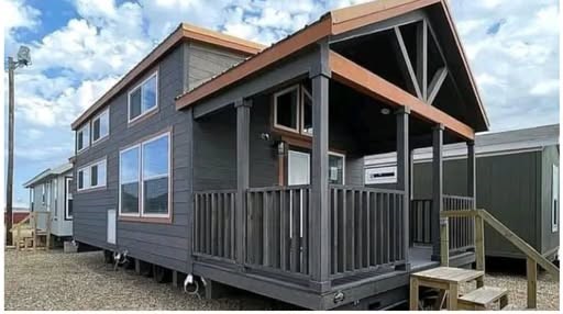 A modern tiny home with dark gray siding and a small porch sits on a gravel lot. Large windows under a cloudy blue sky enhance its cozy feel.