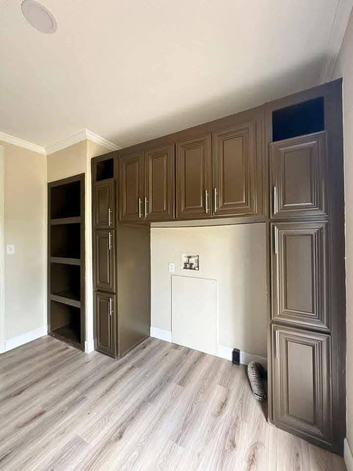 A laundry room with brown wooden cabinets, open shelving, and plumbing connections for appliances. Light wood flooring creates a warm atmosphere.