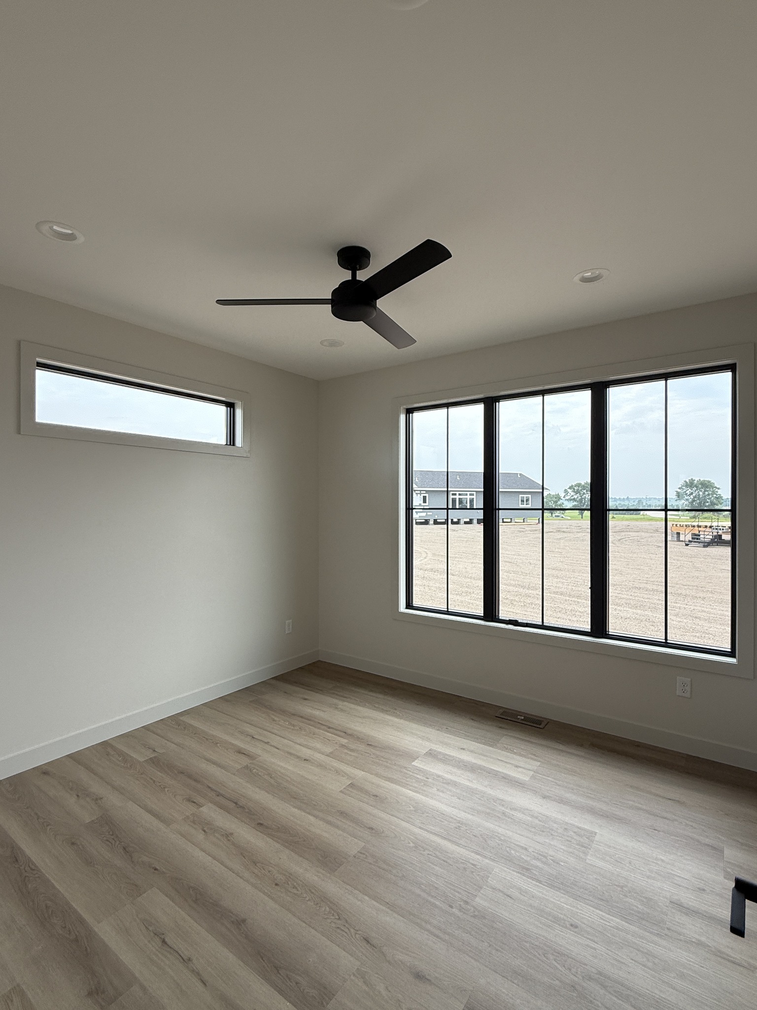 Minimalist room with light wood flooring, white walls, and a modern black ceiling fan. Large window offers a view of a cloudy sky and distant house.