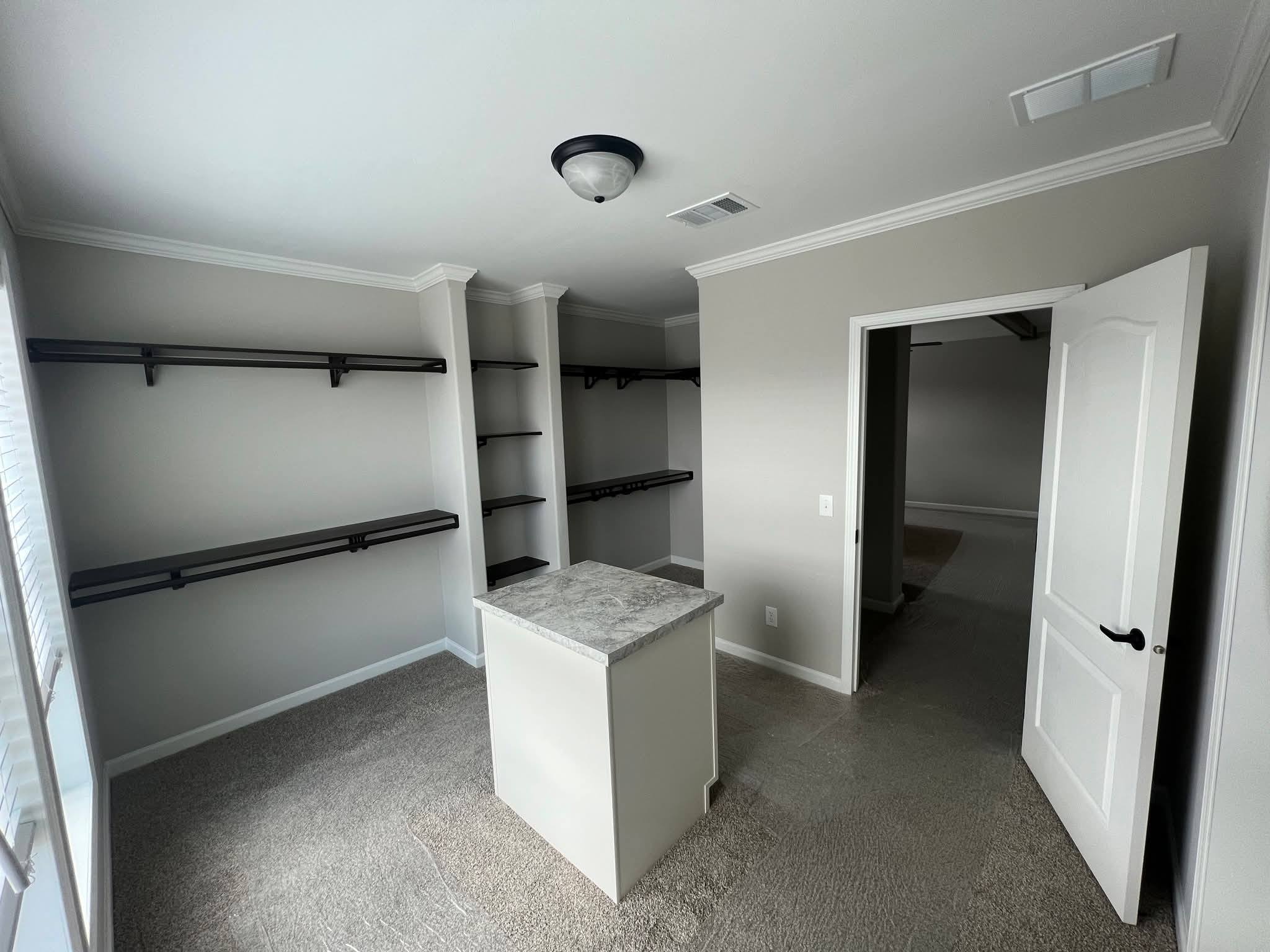Minimalist walk-in closet with gray walls, carpeted floor, and dark wooden shelves. Center island has a marble-like countertop. Open door leads to another room.