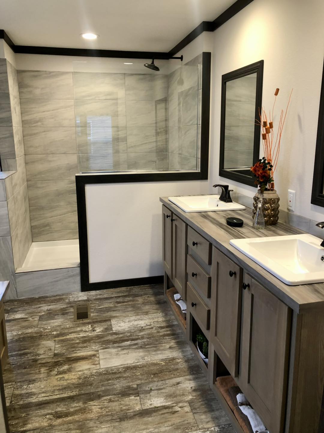 Comprehensive luxury mobile and modular home tour featuring the APH 1662C and premium double-wide floor plans. Exterior highlights include white board-and-batten farmhouse siding and dark charcoal exteriors with rustic wood shutters. High-end interior design features open-concept living rooms with integrated electric fireplaces, shiplap accent walls, and vaulted ceilings with dark wood beams. Gourmet kitchens showcase large marble-look waterfall islands, black industrial hardware, and custom pantry storage with pull-out drawers. The master suites offer bold black accent walls, while luxury bathrooms include freestanding soaking tubs, walk-in tile showers, and LED-mirrored double vanities. Outdoor living is emphasized with a spacious covered front porch featuring a dark wood-plank ceiling and a signature hanging wicker egg chair.