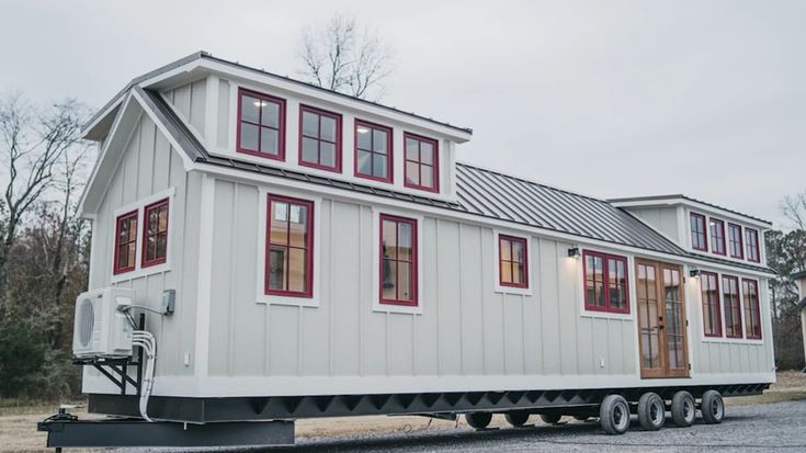 A modern tiny house on wheels with white siding and red-framed windows. It has a gable roof, dormer windows, and a cozy, inviting atmosphere.
