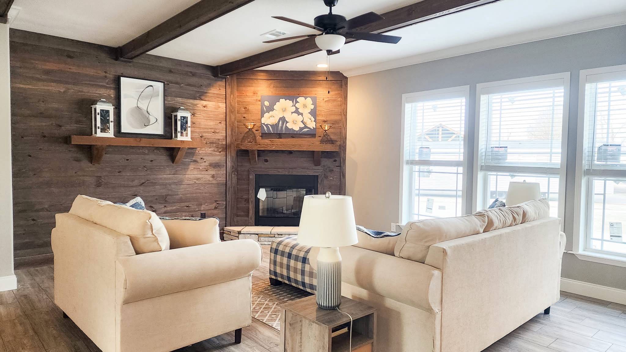A cozy living room with beige sofas facing a wooden fireplace, adorned with artwork and lanterns. Large windows on the right let in natural light.