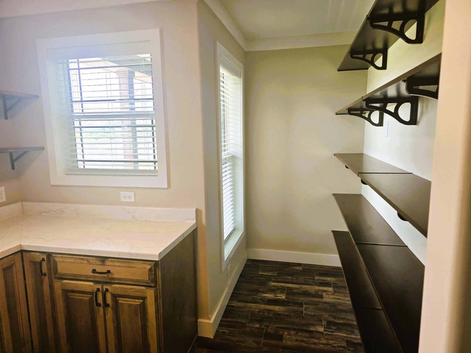 Spacious pantry with wooden shelves on the right, a countertop with wooden cabinets below on the left, and two bright windows letting in natural light.