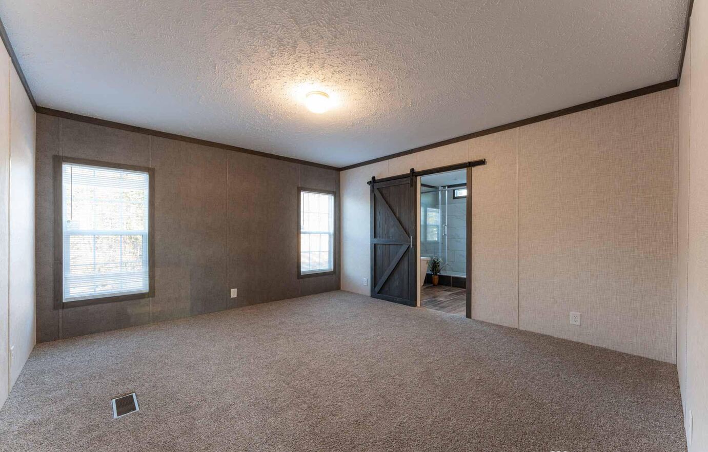 An empty carpeted room with beige walls and two windows. A dark barn door on the right leads to a bathroom. The ambiance is simple and calm.