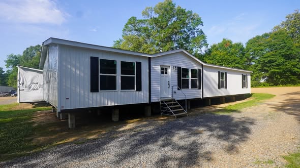 A white manufactured home is elevated on blocks with black shutters and a small staircase at the front entrance. It's set against a backdrop of trees.