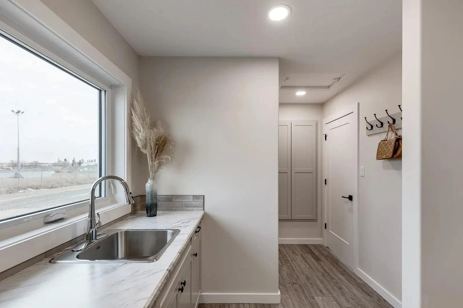 A minimalist utility room with a large window, stainless steel sink, and marbled countertop. Hooks with baskets hang on the wall. Neutral tones convey calmness.