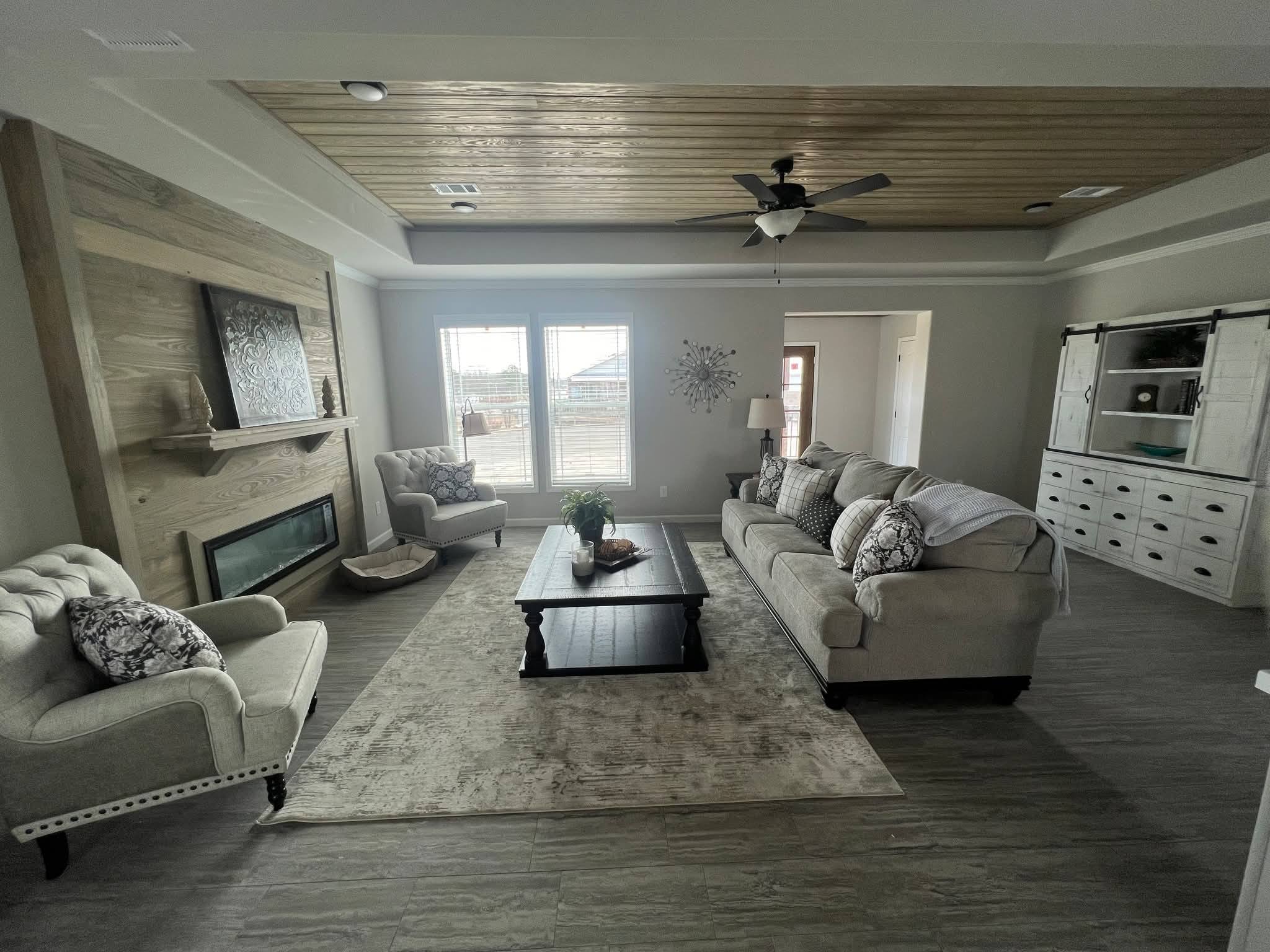 Spacious living room with modern decor, featuring a beige sofa set, wooden coffee table, fireplace, large windows, and a light wood ceiling with a fan.