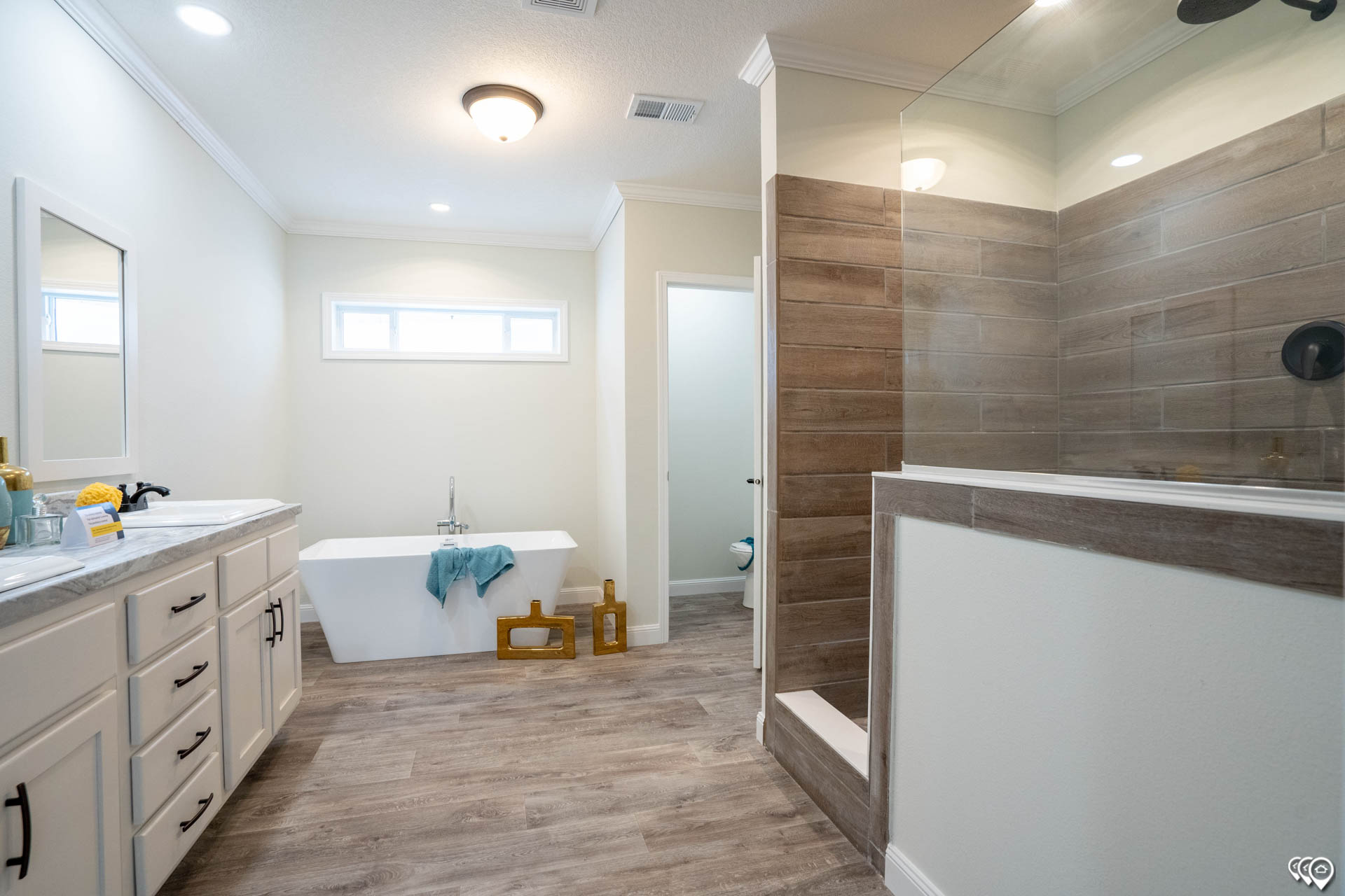Spacious bathroom with a freestanding tub, teal towel, wooden floor, dual-sink vanity, large glass shower, brown tiles, and soft natural light.