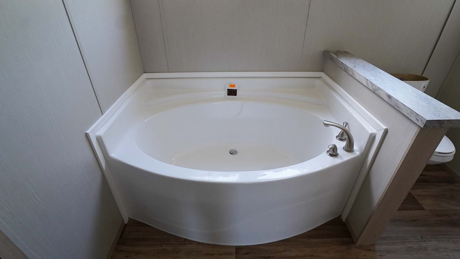 Large white corner bathtub with silver faucet on wooden flooring, set against light gray walls. The setup conveys a clean and modern bathroom design.