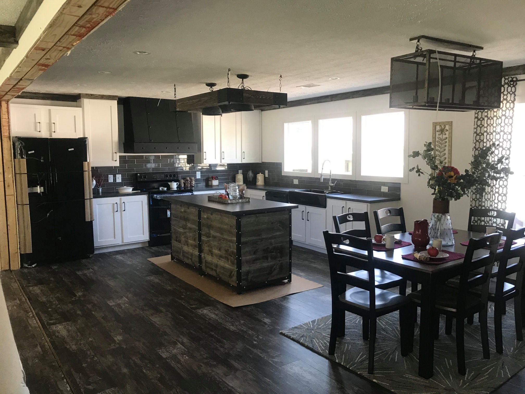 Modern kitchen with dark wood floors, white cabinets, and a central island with a rustic finish. Dining table to the right has a vase with flowers. Bright, inviting atmosphere.