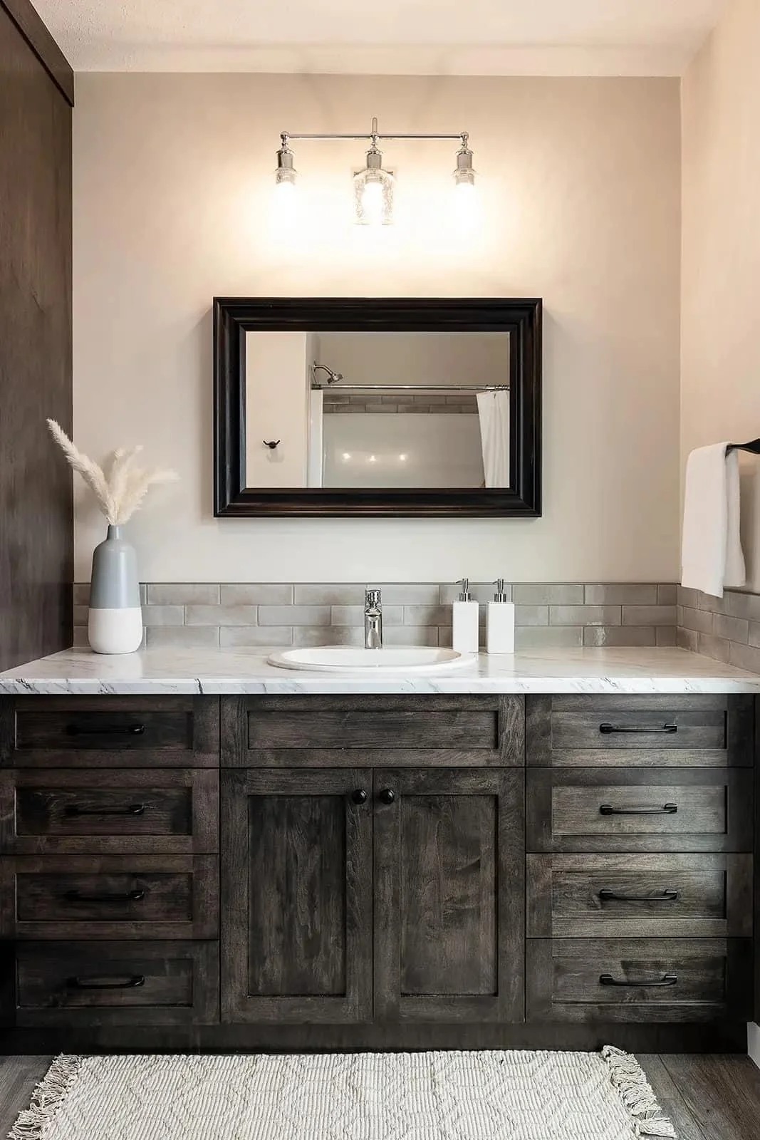 Warm-toned bathroom with a dark wood vanity, light countertop, rectangular mirror, and three-bulb light fixture above. Vase with pampas grass on the left.