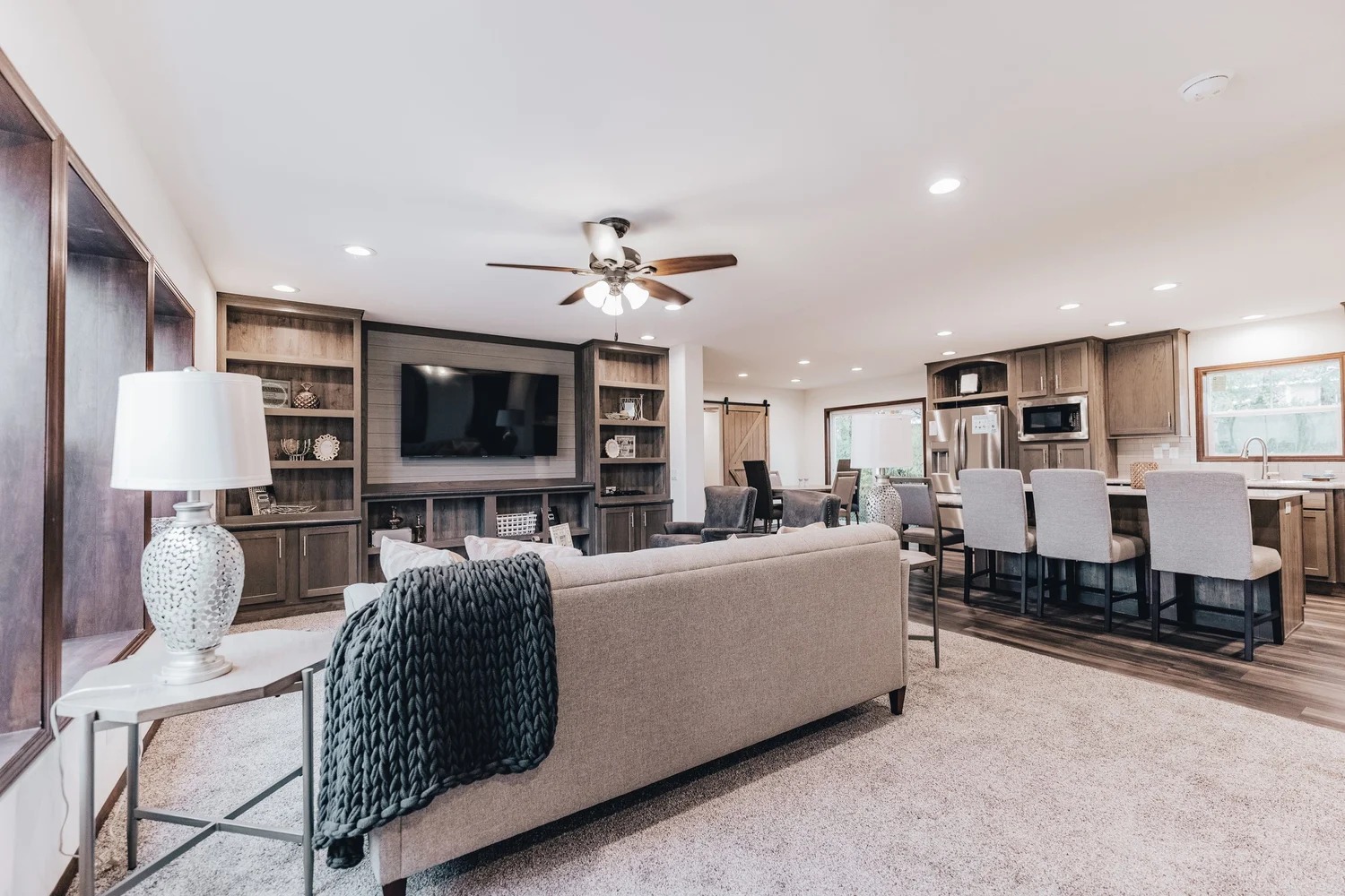 Spacious living room and kitchen with neutral tones. Features a beige sofa, large TV, ceiling fan, built-in shelves, and a kitchen island with bar stools. Cozy and inviting.