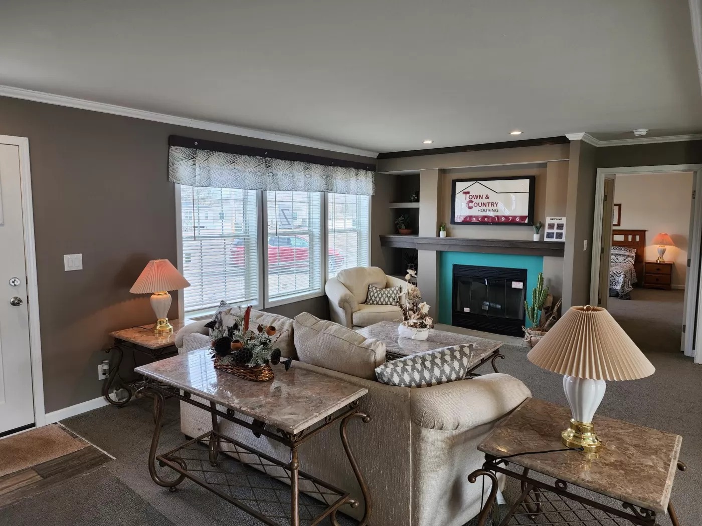 Cozy living room with beige sofas, patterned cushions, and two marble side tables with lamps. A fireplace with mantle is accented by recessed lighting.