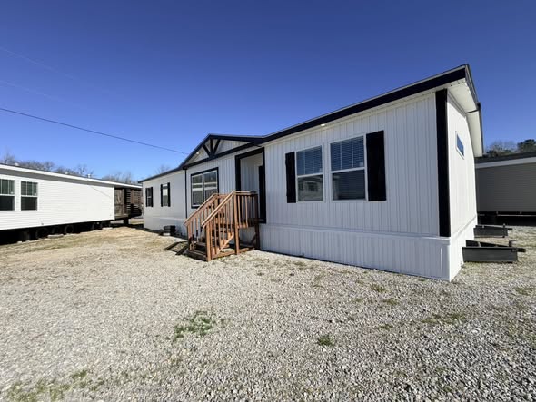 A white mobile home with black trim sits on a gravel lot under a clear blue sky. Wooden steps lead to the entrance, creating a welcoming vibe.