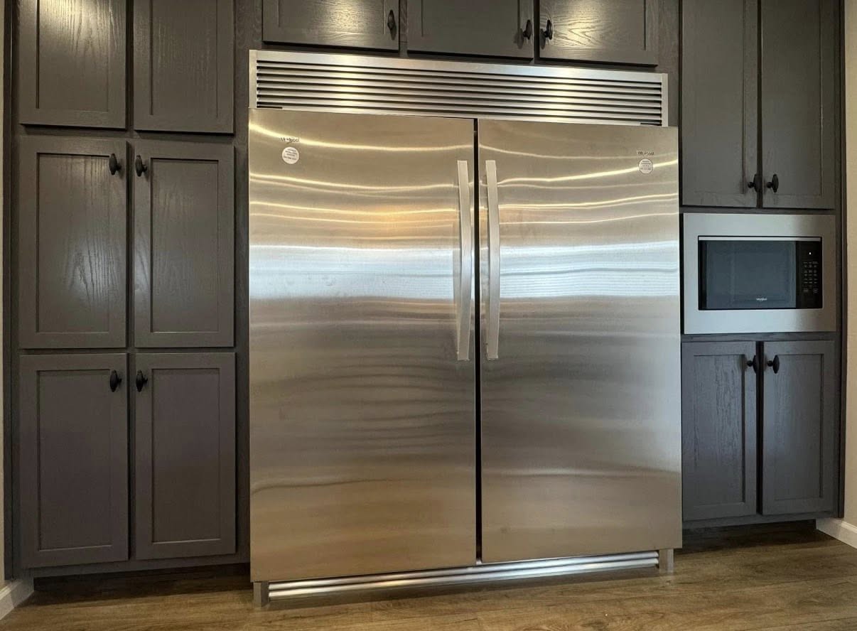 Stainless steel refrigerator with double doors, surrounded by dark wood cabinets with black handles. A microwave is mounted within the cabinet on the right.